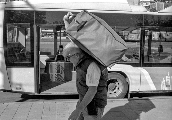 The image depicts a black-and-white street scene captured in front of a bus. In the foreground, an older man wearing sunglasses and a vest is carrying a large, rectangular bag over his shoulder. The man appears to be walking along the sidewalk, while behind him, the bus doors are open and the interior of the bus is visible. The photograph highlights urban life and movement, with the bus and surrounding pavement providing a dynamic backdrop. The gritty texture and high contrast of the photo are characteristic of classic black-and-white film 
