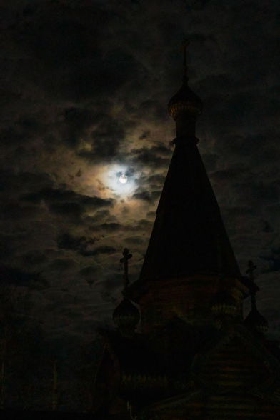 The photo shows the dark silhouette of the Orthodox church's tent-shaped roof, with a full moon shining nearby, illuminating the clouds in the dark evening sky.