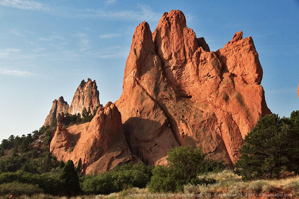 A large jagged rock formation thrusts its way above the ground toward the pale blue sky. Strong contrast between the orange-red rock and the deep green piñon trees around its base.
©BosqueBill.com