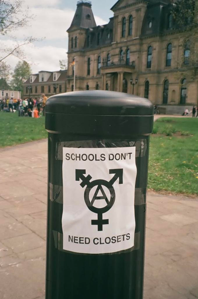 A black and white poster on a small black poll in front of the legislature building says SCHOOLS DONT NEED CLOSETS with a trans anarchy symbol in the middle