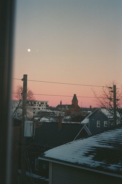 A pink sunset and the moon outside my window with a view of various house rooftops and the city hall building