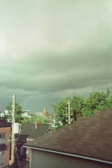 A stormy sky has a faint rainbow behind various rooftops