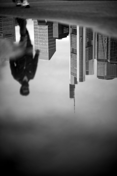 Part of the Frankfurt skyline reflected in a puddle. The blurred silhouette of a passing pedestrian enriches the image.