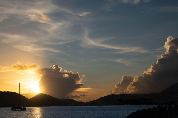 A golden sunset over Charlotte Amalie, St. Thomas, with the sun breaking through clouds above dark silhouetted hills and a lone sailboat drifting on calm water.