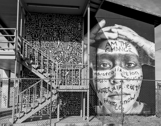 A black and white landscape photo of the end of an abandoned, two-story motel. There's a two-flight staircase on the left that goes up to the walkway on the second floor. The wall behind the staircase is painted in a mosaic of white line geometric shapes on a black background. On the right side of the wall, that is not obscured behind the staircase, is the image of a young Native's face who has a black painted mask across the eyes, like a racoon. Painted on their face are the words, from the forehead down to the chin, "I am change. Industrialization. Pollution. Drought. Water. Air. Earth. Fake Snow. CO2."