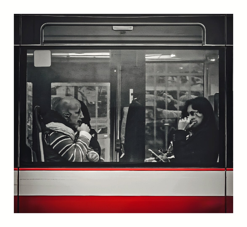This is a photo of a bus interior, through a large window. A bald man with a warped face and striped jacket is sitting on the left. A woman with another warped face and long dark hair is wearing a black top while holding a phone, sits on the right. The bus window has reflections of the outside and interior lights. The bottom part shoes the red and white bus trim.