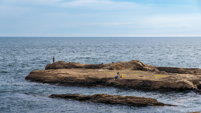 A couple is hanging out on a large rock right by the sea. Another person is fishing.