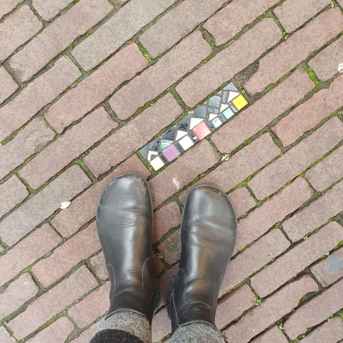 Cobble stones with one of them replaces by a mosaic showing little houses, next to it my feet in black boots. 