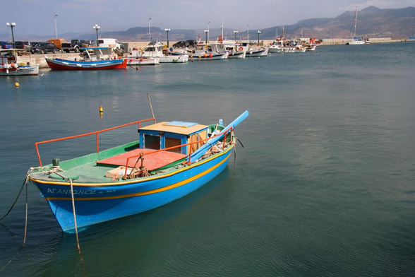 A colorful fishing boat is anchored in calm water at a harbor, with other boats and a distant shoreline visible in the background. The scene features a clear sky and gentle waves.