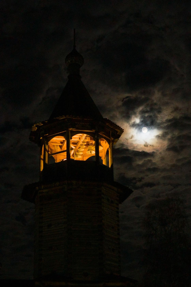The photo shows the top of the bell tower of a wooden Orthodox church. There is a light on the bell tower. A full moon is shining nearby, illuminating the clouds in the darkness.