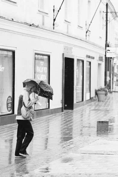 Das Foto zeigt eine Straßenszene in Schwarz-Weiß in der St. Pöltener Innenstadt an einen regnerischen windigen Tag. Man sieht eine Person mit Regenschirm der durch den Wind verbogen ist.