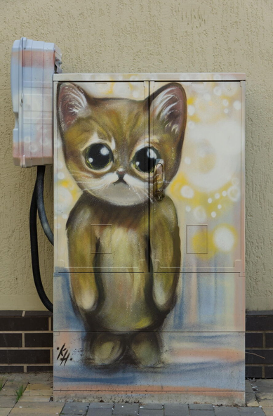 In the image, there is a large electrical box painted with an adorable cat image. The painting covers the entirety of the electrical box, making it appear as if the cat is standing in front of a door or window. The cat is depicted in shades of brown and white, standing upright on its hind legs, exuding an air of curiosity and intrigue with its large dark-green eyes staring directly into the camera. Its nose is colored in shades of pink and orange, adding to its charm. The background of the mural is a mix of yellow and pink colors, creating a warm and inviting atmosphere. The artist's signature "KH" is discreetly placed in the bottom left corner of the painting, marking their creative territory on this urban canvas. The medium used for this piece appears to be spray paint, given the slightly rough texture visible throughout.
