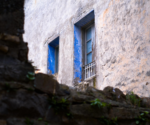 Fotografía de unas ventanas tras un muro de piedra ruinoso. Las ventanas tienen el marco interior azul. La pintura y fachada estan curtidas por el tiempo y los elementos con texturas interesantes