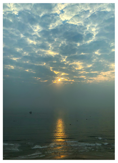 Photo of a beach in the early morning, taken from a high vantage point. The sun pierces through a dramatic cloud-filled sky and is reflected by the sea in a glittering column that stretches to the shore. Distant smog softens the horizon so that it’s hard to tell where the sea ends and the sky begins. Tiny human figures dot the sand and wade into the water to meet foam-edged waves rolling ashore. Tiny boats are scattered here and there on the sea.