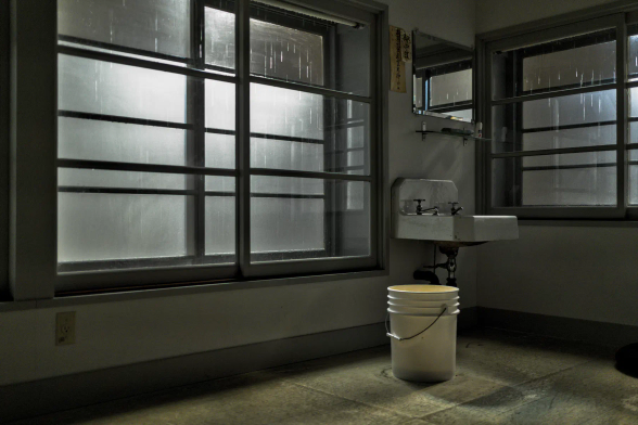 Blick in einen karg eingerichteten Raum. Zwei Wände treffen sich in einer Ecke. Links ein breites und recht tiefes Fenster, aufgeteilt mit sechs Sprossen, daneben ein einfaches Waschbecken mit zwei Wasserhähnen und einem einfachen Spiegel darüber. Rechts schließt sich das zweite Fenster an, etwas schmaler aber auch aufgeteilt mit sechs Sprossen. Ein Eimer steht auf dem Boden. Die Wände sind schmucklos, nur neben dem Spiegel hängen zwei Zettel mit japanischer Schrift. Hinter den Fenster ist zu sehen, wie der Regen fällt. Kaltes, fahles Licht fällt durch die Fenster in den Raum. 
.
A view into a sparsely furnished room. Two walls meet in a corner. On the left is a wide and quite deep window, divided by six panes, next to it a simple washbasin with two taps and a plain mirror above it. On the right is the second window, slightly narrower but also divided by six panes. A bucket stands on the floor. The walls are bare, except for two pieces of paper with Japanese writing hanging next to the mirror. Behind the windows, rain can be seen falling. Cold, pale light falls through the windows into the room. 
.