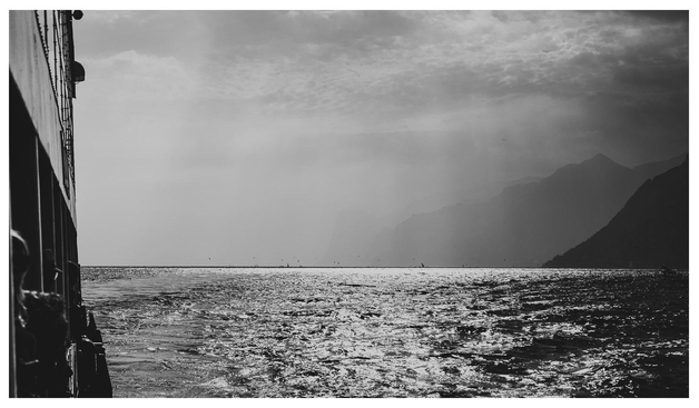Schwarz-Weiß Bild von einem Schiff in Richtung Sonne geschossen. Das Wasser glänzt, Berge im Hintergrund sind im Dunst geschichtet. 