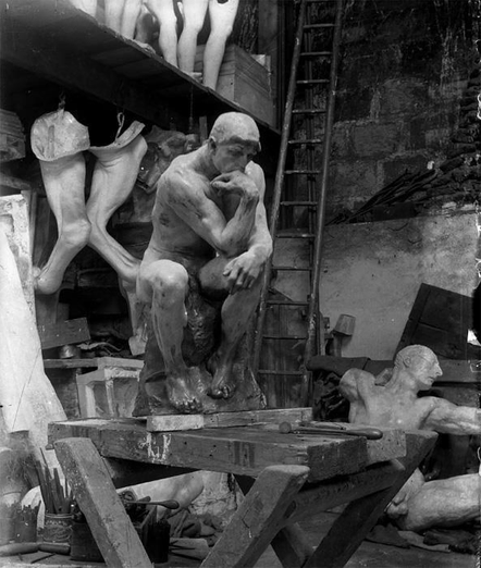Photo en noir et blanc de l'intérieur d'un atelier de sculpture, une reproduciton du penseur de Rodin est posé sur un établi, des bouts de corps sont posés et accrochés autour de lui dont posé en bas à droite l’œuvre de Bourdelle, Heracles