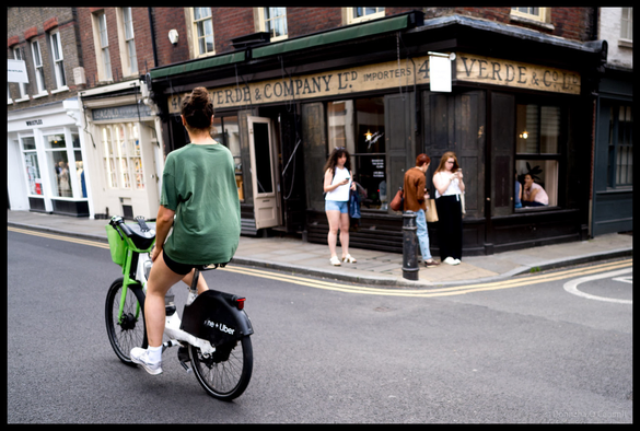 A woman in green t-shirt on a bright-green bike rides past historic black-painted corner shop with gold lettering reading "VERDE & COMPANY LTD IMPORTERS" on a London street, with pedestrians visible near shopfront and period brick buildings in background.