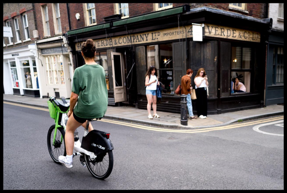 A woman in green t-shirt on a bright-green bike rides past historic black-painted corner shop with gold lettering reading "VERDE & COMPANY LTD IMPORTERS" on a London street, with pedestrians visible near shopfront and period brick buildings in background.
