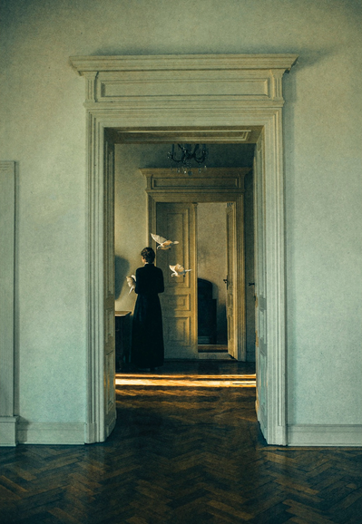 A photograph taken from a room with a herringbone-patterned wooden floor, looking through an ornate doorway into another room. Inside the second room, a person in a long, dark dress stands with their back to the viewer, facing a closed door. Three white doves are in mid-flight around them, illuminated by a bright sliver of light coming from under the door.