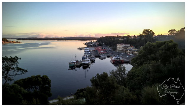 A tranquil, high angle view of the Strahan waterfront at sunset, looking across Macquarie Harbour. The sky features a soft, pale blue transitioning to golden pink light just above the distant tree lined horizon.  The water is still and dark, reflecting the colours of the sky. In the middle ground, a cluster of fishing boats and small commercial vessels are tied up at a pier, adjacent to historic town buildings nestled among dense greenery.  Dark foliage frames the bottom and right side of the photograph.