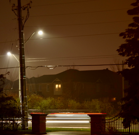 Blurry lines go by from a car’s red and head-lights at night on a street, and above with electrical wires.

A residential metal fence is attached to two brick posts, opening to a sidewalk.

There are three tall, lit lampposts and some houses with lights on. Trees and bushes are on each side of the street.

There is also a slight fog in the night's air after a mild evening rainfall.