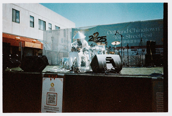 a scan of a color film photograph of a lone guitar player sitting on stage dressed in silver everything, including a silver cone hat. in the background, the text says oakland chinatown street festival