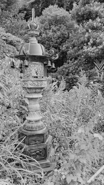 Eine große Lampe aus Stein mit einem Kupferdach, an welchem mehrere Glöckchen hängen. Die Lampe hat runde und sechseckige Elemente in geschichteten Stufen. Umgeben wird die Lampe von Grün. Schwarz-weiße Aufnahme.