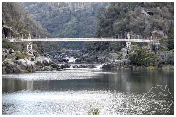 A photograph signed by Kev Peirce showing the Alexandra Suspension Bridge across the Cataract River in Launceston's Cataract Gorge.  The white, truss style bridge spans the water, supported by white towers on either bank. A wide, still section of the river is in the foreground, leading to rocky rapids and a small weir beneath the bridge.  The steep banks and hillsides are covered in dense, dark green and grey-brown Australian bushland.