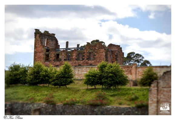 Ruins of the Port Arthur Penitentiary, a large brick structure with sections of the roof and upper walls missing, standing behind a lower, intact brick wall.

The foreground features a grassy mound with small green trees and some red flowered shrubs. The sky above is cloudy with patches of blue.