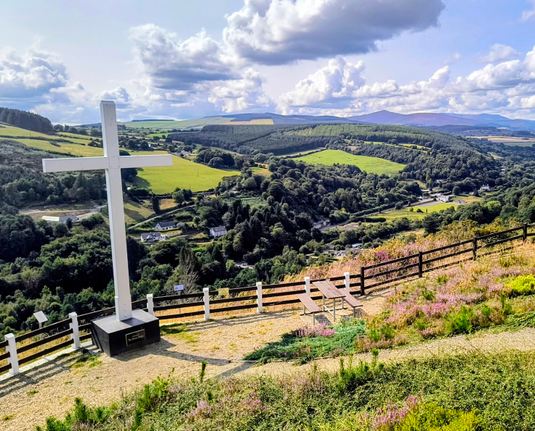 Von einer Erhöhung aus hat man einen Blick in ein Tal und über die sonnigen Wicklow Berge mit Nadelwald und hellgrünen Wiesen. Am Himmel sieht man Wolkenbänder. Im Vordergrund ist ein Zaun, welcher den hellen Schotterplatz umgibt, dort stehen Picknickbänke. Auf der linken Seite steht ein weißes Kreuz auf einem Sockel, es ragt bis in den Himmel hinein. Im Vordergrund wächst neben vielen Grün, rosa Heide.
