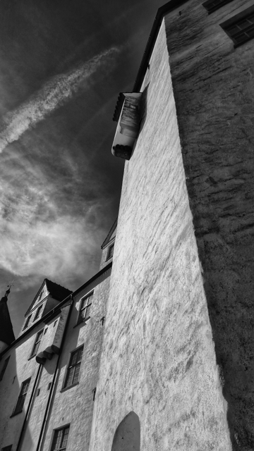 Eine Schwarzweißfotografie, die von unten nach oben entlang der Fassade eines Schlosses aufgenommen wurde. Rechts im Bild ist ein hoher Turm zu sehen, links ein niedrigerer Bau mit Fenstern und Dacherkern. Der Himmel ist leicht bewölkt. 

A black-and-white photograph taken from the bottom to the top along the façade of a castle. On the right side of the image, a tall tower can be seen, and on the left side, a lower building with windows and dormers. The sky is slightly cloudy. 