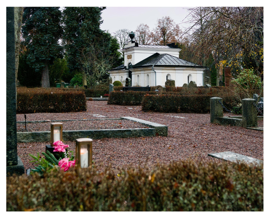 En liggande bild på en kyrkogård med en byggnad i vit puts, från början en vintergravplats numera förråd. I förgrunden grusgångar, bruna häckar och längst fram rosa blommor med ljus på en grav. Träd i bakgrunden.
