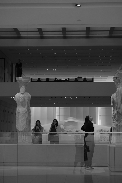 Black+white photograph of the Carytids in the Acropolis Museum, Athens. The architecture of the museum with mezzanine levels and lit ceilings is visible. A glass wall creates a layer of reflections at the floor level.  Two Carytids are visible from behind, graceful and commanding. Two women are viewing one of them and taking photos with their phone. Another woman is viewing the other, gazing up at it with her hands on her hips. Acropolis Museum, Athens, Greece, 27 November 2019. Photo Cindy Kohtala