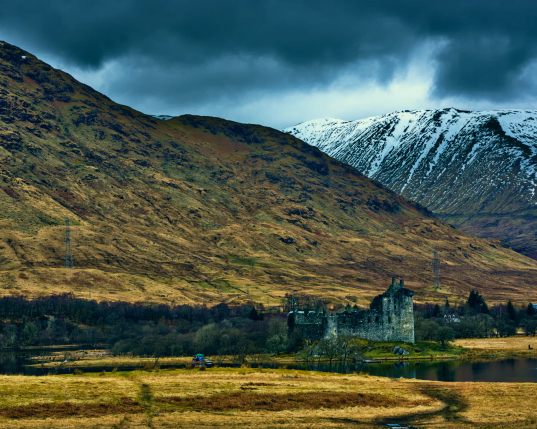 In Vordergrund eine Wiese, noch braungelb vom Winter- In der Bildmitte erhebt sich Klichurn Castle an einem kleinen See. Dahinter erhebn sich Berge, einige noch braungelb vom Winter, rechts ein Berg, der noch schneebedeckt ist. Darüber rollen dunkle Regenwolken über das Tal. 