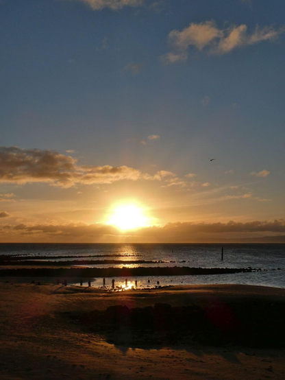 MId-november sun setting over the sea, about to descend into a band of cloud over the horizon. The sky is already very dark but you can tell it is blue with a few clouds. The foreground is pitch black with some outlines of rock patches and poles. The sea reflects the setting sun.