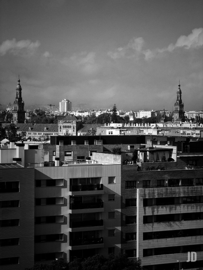 Una fotografía en blanco y negro de un paisaje urbano, mostrando una mezcla de arquitectura moderna y clásica bajo un cielo parcialmente nublado. En primer plano, varios edificios residenciales modernos de múltiples plantas con balcones y ventanas proyectan fuertes sombras. Detrás de ellos, en el medio plano, se distingue un edificio más antiguo y ornamentado con un tejado distintivo. En el fondo, dos imponentes torres con chapiteles puntiagudos se elevan sobre el horizonte de la ciudad, una a cada lado de la imagen, con una densa extensión de edificios urbanos y algunas colinas o montañas apenas visibles en la distancia. El alto contraste de la imagen resalta las formas y texturas.