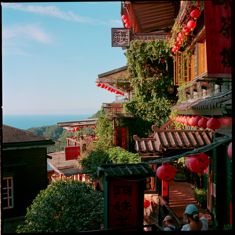 A narrow, vine-covered alleyway lined with traditional wooden buildings adorned with red lanterns and Chinese characters. The scene overlooks a distant ocean under a clear blue sky, blending cultural architecture with natural beauty.