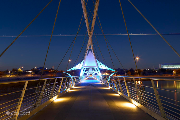 This is an abstract colour sunset photo in landscape format looking along a pedestrian bridge. Tempe, Arizona (2012).

The camera is aligned with the centre of a modern pedestrian bridge, with footlights lighting the ground, spaced every few metres. Along either side are two metal railings angled inwards about thirty degrees from the vertical. These railings converge together with increasing distance so as to nearly touch at the end of the bridge, perhaps a hundred metres away. About ten metres away are two white tubular, near vertical frames forming an 'X' with stout rods descending downwards to the bridge in the fashion of a cable-stayed suspension bridge. Just beyond the support is an illuminated awning stretched out in a sharp, pointed triangle. A second 'X' frame can just be made out in the distance. On the right side of the bridge is a still, body of water. On the left side - darkness. Above our bridge is a clear and cloudless blue sky, slightly orange along the ground but rapidly graduating into blue and becoming darker towards the top of the image. About two-thirds up in the image is a straight, bright line stretching across the frame from left to right, with pairs of white dots of lights evenly spaced above the line, while on the line are occasional red dots of light. 

The bridge is the Tempe Dam Pedestrian & Bike Bridge, with the Tempe Town Lake on the right of the photo. The line of light is from an aircraft landing a Phoenix airport. 