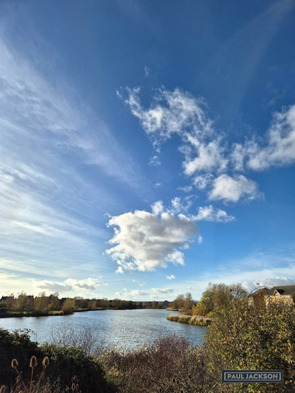 A bright, crisp autumn scene by a  lake, dominated by the vast, bright blue sky and its dynamic cloud formations. The upper atmosphere features long, delicate streaks of cirrus clouds running horizontally and diagonally across the frame, suggesting high-altitude winds and lending a sense of motion to the scene. These wispy formations contrast sharply with the intense blue, creating texture and depth.

Below these high clouds sits the central feature: a spectacular, puffy cumulus cloud. This singular white mass is brightly illuminated by the sun, showcasing three-dimensional form and casting subtle shadows on itself. This vibrant contrast between the high, streaked clouds and the solid, brightly lit lower cloud anchors the sky, drawing the eye upward before it settles on the peaceful lake and autumn landscape below.

The strong daylight illuminates the water's surface, which remains dark and relatively still, bordered by low, autumnal trees and shrubs displaying muted browns and dry greens.

The foreground is dominated by natural, overgrown foliage, including tall, dry reeds and bushes, which frame the reflective water stretching into the distance. Houses on the far bank peak through the sparse trees, indicating a scenic but accessible walking area on a beautiful November day.

In one corner of the image, there's the photographers name that reads "PAUL JACKSON" in white text on a dark rectangular background.