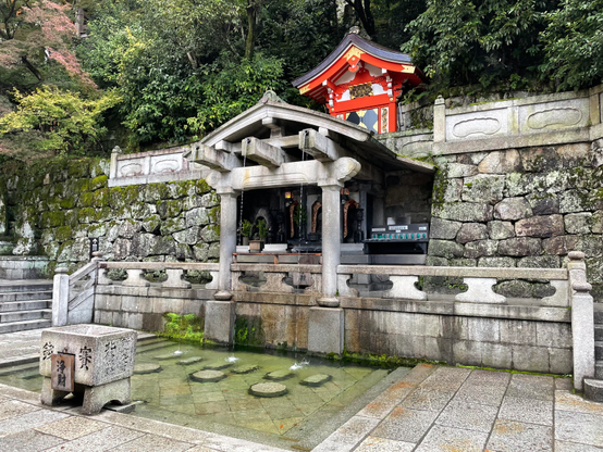One of the shrines in Kiyomizu-dera. A stone wall and adjoining fountain set a peaceful, centering aura, while a mini red temple sits atop the structure, requesting reverence and tranquility. In the background are many trees, some of which are transitioning to red. 