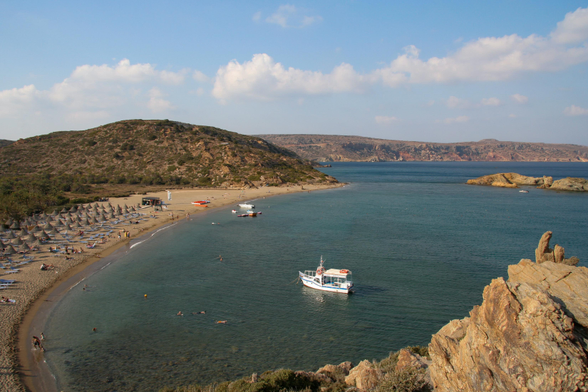 A scenic beach view featuring white sand, umbrellas, and sun loungers along the shore. Clear blue waters with a few boats, and people swimming and relaxing. A hilly coastline and a few clouds in the sky complete the tranquil setting.