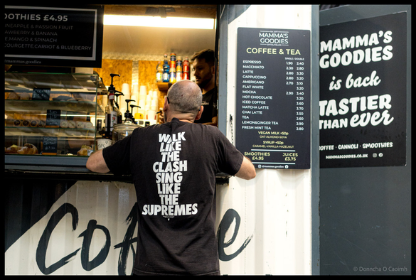 Customer in black t-shirt with white text reading "Walk Like The Clash Sing Like The Supremes" ordering at Mamma's Goodies coffee kiosk counter at Spitalfields Market London, with menu boards showing coffee and tea prices visible and barista working inside brightly lit service window.