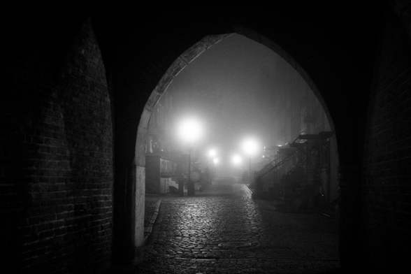 A foggy street at night in Gdansk, near Motława river. The picture is taken through the arched vault from the riverbank promenade. Old-fashioned streelamps illuminate an empty, narrow cobblestone street. Hints of buildings can be seen by them in the fog. The buildings are very old in style, but most of Gdańsk was re-built after WWII so it’s anyone’s guess whether they really are older than that. (If locals read this, maybe they can share more info on the subject?) The end of the street cannot be seen, it vanishes into fog. The street in question might be Mariacka, but I’m not sure.