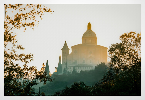 This is a photo of a sunlit, misty hilltop featuring a grand, dome-topped building with a smaller, pointed spire to the left (Il Santuario della Madonna di San Luca). The architecture is yellow/brown stone with ornate details. The surrounding trees frame the scene. They have dark green foliage contrasting with the golden sunlight. The sky is clear, casting a warm evening glow.