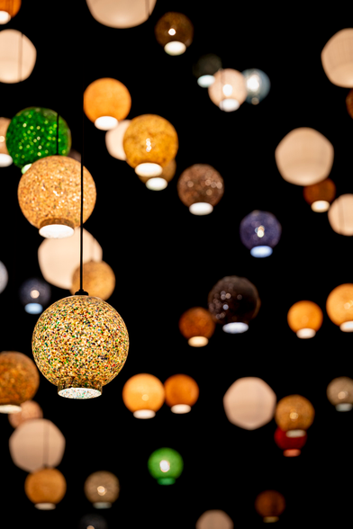 Heel veel ronde lampenkappen van verschillende kleuren. Ze hangen op verschillende afstanden en verschillende hoogte voor een zwarte achtergrond.