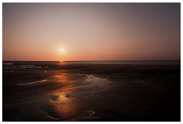 In the foreground, a wet beach illuminated by the setting sun can be seen. In the background, the sea. A lone angler stands on a pier.