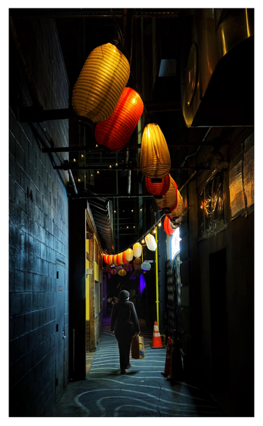 Nighttime photo of a narrow alley illuminated by hanging Japanese lanterns in alternating colors of red and yellow. The gray pavement is marked with wavy white stripes. A person in a dark winter hat and coat stands with their back to the camera in the middle distance, carrying a paper shopping bag and illuminated by the light from a restaurant storefront on the right side of the alley.