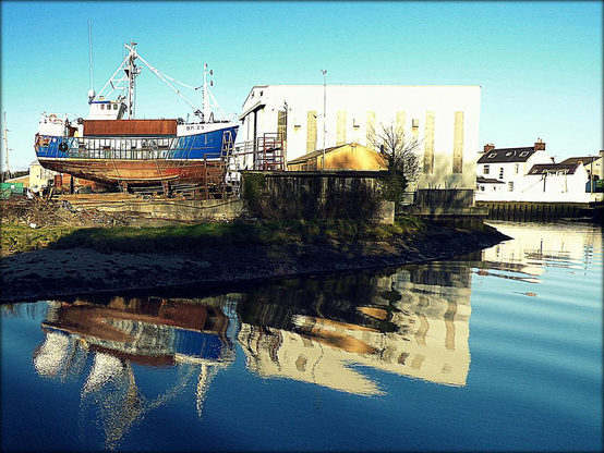 A colour photograph showing a boat in dry dock in Noble's boatyard, Girvan . The blue trawler, contruction hall and deep blue afternoon sky above are mirrored in reflection in the wateter of the river Girvan spliting the frame of the shot.