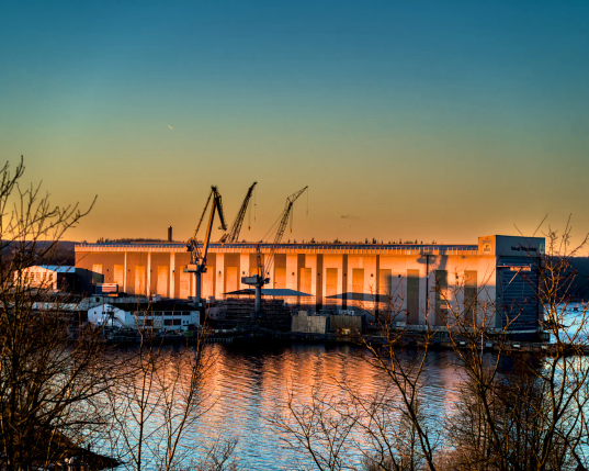 Von einer kleinen Anhöhe herab sind die Flensburger Förde und die Flensburger Schiffswerft im Abendlicht zu sehen. Die große Werfthalle leuchtet im goldenen Licht der untergehenden Sonne. Der Himmel über der Werft verfärbt sich von Rotgold über blau zu dunkelblau. Ein Schwarm Vögel zieht über die Werft. 
.
From a small hill, the Flensburg Fjord and the Flensburg shipyard can be seen in the evening light. The large shipyard hall glows in the golden light of the setting sun. The sky above the shipyard changes colour from reddish gold to blue to dark blue. A flock of birds flies over the shipyard. 
.
